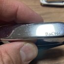 Detailní pohled na kovový, chromovaný předávej nebo kryt s vyhlacenými okraji. Na povrchu je vyryt nápis BaCH s malým symbolem blesku na konci. Povrc…