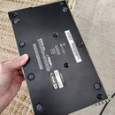Fotografie zachycuje spodní stranu černého elektronického zařízení s kovovým rámem v zlatavé barvě. Na spodní části jsou vidět gumové nožičky a někol…