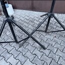 Dvě černé kovové stativy s trojnožkami umístěné na dlažbové ploše. Povrch laku je v obou případech výrazně opotřebovaný s četnými odřapkáním a škrába…
