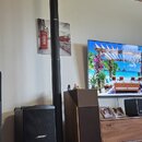 Na fotografii jsou dva černé reproduktory značky BOSE, z nichž jeden je vysoký sloup a druhý je subwoofer s mřížkou. Vedle nich stojí dřevěný reprodu…