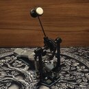 Černý bicí pedál s kovovou konstrukcí a stříbrným šlapadlem. Mechanismus pohonu je řešen řetězem a ozubeným kolem. Na zařízení nejsou vidět žádné zna…