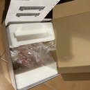 Otevřená krabice s bílým polystyrenovým vložkem, ve které se nachází průhledný plastový sáček s příslušenstvím. Nad krabicí je položen papírový návod…
