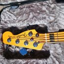 Detail hlavy basové kytary v barvě žlutého javoru, umístěné v polstrovaném kufru. Na hlavě je černý nápis Fender Jazz BASS. Jsou vidět čtyři kovové l…