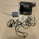Na fotografii je černý napájecí adaptér s nápisy ADAM, CIOKS a textem "NOT HOT PLUG". K adaptéru patří černý napájecí kabel a sada různých propojovac…
