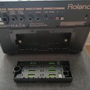 Zadní strana černého komba značky Roland. Jsou vidět konektory DC IN, FOOT SW, LINE OUT a AUX IN s doprovodnými popisky funkcí. V dolní části je vyjm…