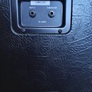 Detail zadní strany černého kytarového boxu s texturovaným povrchem. V horní části je ovládací panel se dvěma vstupy označenmi INPUT a THROUGH, nápis…