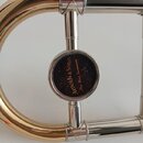 Detailní pohled na část mosazného a niklovaného dechového nástroje. Uprostřed je kulatý černý štitek s nápisem „Arnolds & Sons® Fine Wind Instruments…