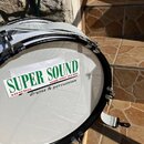 Bílý buben s černým obručováním a kovovými šroubnicemi. Na bláně je nalepený bílý papírový štítek s nápisy SUPER SOUND, mobil: 0905 194 665, 949 01Ni…