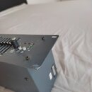Šedý kovový box s ovládacím panelem, který obsahuje posuvníky, otočný knoflík a kontrolky. Na panelu jsou viditelné nápisy INPUT GAIN, LOW OUT a HI O…