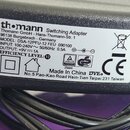 Detailní záběr na štítek černého spínaného adaptéru. Na štítku je uveden text: th•mann Switching Adapter, Thomann GmbH - Hans-Thomann-Str. 1, 96138 B…