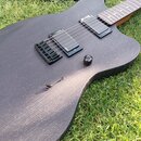 Černá elektrická kytara s matným povrchem a viditnou strukturou dřeva, položená na trávě. Nástroj má dva černé humbuckery s bílým nápisem emg, jeden…