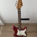 červená elektrická kytara s bílým pickguardem a hnedým hmatníkem. Na hlavě je černý nápis Squier STRATOCASTER by Fender. Nástroj stojí na černém stoj…