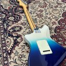 Zadní strana elektrické kytary s barevným přechodem od bílé přes modrou až po tmavě modrou. Na zadní straně krku je vidět nápis Fender. Tělo má v hor…