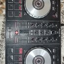 Černý DJ kontrolér Pioneer DJ DDJ-SB2 s logem serato v levém dolním rohu. Zařízení má dva stříbrné jog-wheels, fadery a řadu ovládacích tlačítek s če…
