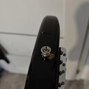 Detailní pohled na černý, lesklý hриф hudebního nástroje. Je vidět jeden kovový ladič s vložkou pro imbusový klíč, který vykazuje známky koroze a opo…
