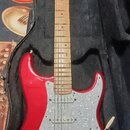 Červená elektrická kytara typu Stratocaster s bílým perleťovým pickguardem, umístěná v černém softcasu. Na hlavě hmatníku je nápis Fender Stratocaste…