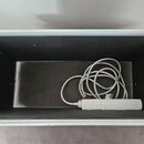 Hliníkový přepravní kufr s černým vnitřním povrchem. Uvnitř leží bílý svitící tubus s připojeným bílým napájecím kabelem. Na bočních stěnách jsou vid…