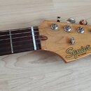 Hmatník a hlava elektrické kytary z přírodního dřeva s tmavým hmatníkem. Na hlavě je černý nápis Squier by Fender a STRATOCASTER®. Jsou vidět šest la…
