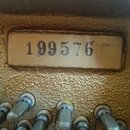 Detailní záběr na část hudebního nástroje se zlatým povrchem. Na obdélníkovém štítku je černý nápis 199576. Ve spodní části jsou vidět kovové kolíky…