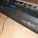 Černý digitální klavír s nápisy YAMAHA a PSR-S670. Na levé straně jsou vidět ovládací prvky, displej a několik vstupů, do kterého je zapojen černý ka…