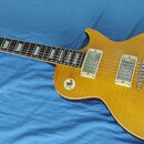Žlutá elektrická kytara typu Les Paul s hmatníkem z tmavého dřeva a trapezoidními vložkami. Na hlavě je nápis Gibson. Kytara má dva humbuckery s kryt…