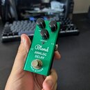 Zelený efekt pedál s kovovým povrchem. Na přední straně je bílý nápis „Blond ANALOG DELAY“ a v dolní části text „TRUE BYPASS“. Pedál má tři černé ovl…