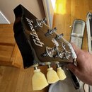 Detail hlavy černé elektrické kytary s bílým nápisem Gibson. Na hlavě jsou instalovány šest kovových ladících kolíků s plastovými světlými knoflíky.…