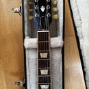 Černá elektrická kytara v otevřeném kufru s bílou vnitřní výplní. Na hlavě hmatníku je bílý nápis Gibson a logo v podobě koruny. Hmatník má tmavé dře…