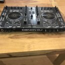Černý DJ kontrolér s dvěma jog-koly, faderem a řadou ovládacích knoflíků a tlačítek. Na spodní hraně je bílý nápis DENON DJ. Zařízení leží na dřevěné…