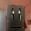 Detail zadní strany elektrické kytary s tmavě červeným lakem. Na těle je přišroubován černý plastový kryt s dvěma svislými výřezy, skrze které jsou v…