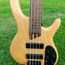 Světle hnědá elektrická basová kytara s přírodním dřevěným povrchem a hnědým hmatníkem. Nástroj má dva černé humbuckery, tři černé ovládací knoflíky…