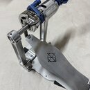 Detailní pohled na kovový pedál basového bubnu v stříbrné barvě s modrými prvky. Na šlapce je vyryto logo znázornující kříž v čtverci. Na horním mech…