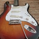 Elektrická kytara s trupem v barevnosti sunburst a bílým pickguardem. Na pickguardu je vidět tři snímače s bílými plastovými krytkami a nápis "MADE I…