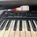 Detailní pohled na ovládací panel a klávesnici elektronického nástroje. Na černém panelu je bílý nápis Organ/Strings O9 a RS-09. Horní část obsahuje…