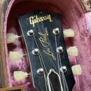 Detail hlavy černé elektrické kytary značky Gibson, model Les Paul, v otevřeném pouzdře s růžovým polstrováním. Na hlavě je zlatý nápis Gibson a kurz…