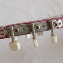 Detail hlavy červené kytary s kovými ladicími mechanismy a bílými plastovými knoflíky. Kovové kryty mechanismů jsou zdobeny rytým ornamentem. Povrch…