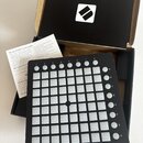 Černý MIDI kontrolér Novation Launchpad Mini s mřížkou bílých podsvícených políček. Na spodní liště jsou nápisy novation a LAUNCHPAD MINI, vpravo je…