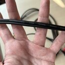 Černý audio kabel s nápisem PROFESSIONAL LOW NOISE AUDIO CABLE. Kabel je z pružného materiálu a drží ho lidská ruka. V pozadí jsou vidět další části…