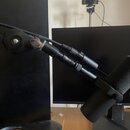 Černý mikrofon s nasazenou pěnovou windsockem, umístěný v černém rameni mikrofonního stativu. Na spodní části monitoru v pozadí je vidět nápis msi. P…