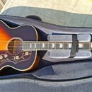 Akustická kytara s korpusem v barvě sunburst, položená v šedém polstrováním pouzdře. Na hmatníku jsou vidět perleťové vložky a na horní dece pod žstř…