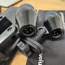 Dvě černá dynamická mikrofony s mřížkou, umístěná v držácích. Na spodní části obou mikrofonů je nápis MADE IN GERMANY. Pod nimi leží černý textilní o…