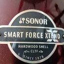Detailní záběr na kovový štítek připevněný k tmavě červenému povrchu nástroje. Na štítku je uveden nápis SONOR, SMART FORCE XTEND, HARDWOOD SHELL, CL…