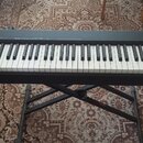 Černý klavírní kontrolér značky Roland, model A-88. Nástroj je umístěn na černém X-stojanu. Na levé straně jsou ovládací prvky, knofíky a tlačítka. P…