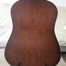 Zadní strana akustické kytary v hnědé barvě s viditnou strukturou dřeva. Kytara má světlý lem po obvodu korpusu a stojí na černém stojanu. Na fotogra…