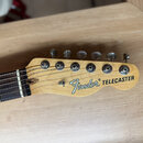 Detail hlavy elektrické kytary z přírodního světlého dřeva. Na hlavě je černý nápis Fender® TELECASTER a pět kovových ladiček. Hmatník je z tmavého d…