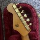 Zadní strana hlavy kytary z přírodního dřeva v červeném polstrovaném kufru. Na hlavě jsou zlaté ladicí kolíky a nápis Designed and Backed by Fender v…