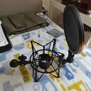 Černý pop-filtr s kulatou akustickou clonou a kovovým rámem v podobě klece. Filtr je vybaven šroubovacím uchopovadlem pro upevnění na stojan. V pozad…