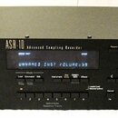 Černý rackový sampler s nápisy ensoniq, ASR-10 a Advanced Sampling Recorder. Přístroj má digitální displej s modrým textem UNNAMED INST VOLUME=99 a S…