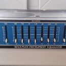 Černý rackový grafický ekvalizér s modrým panelem a bílými posuvníky. Na zařízení jsou nápisy PREAMP, BASS BAND, MID BAND, TREBLE BAND a OUTPUT, v do…