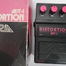Černý kytarový efekt s nápisem DISTORTION ADT-1 a třemi růžovými oválnými knoflíky s popisky LEVEL, TONE a DIST. Pod efektem je černý plastový kryt s…